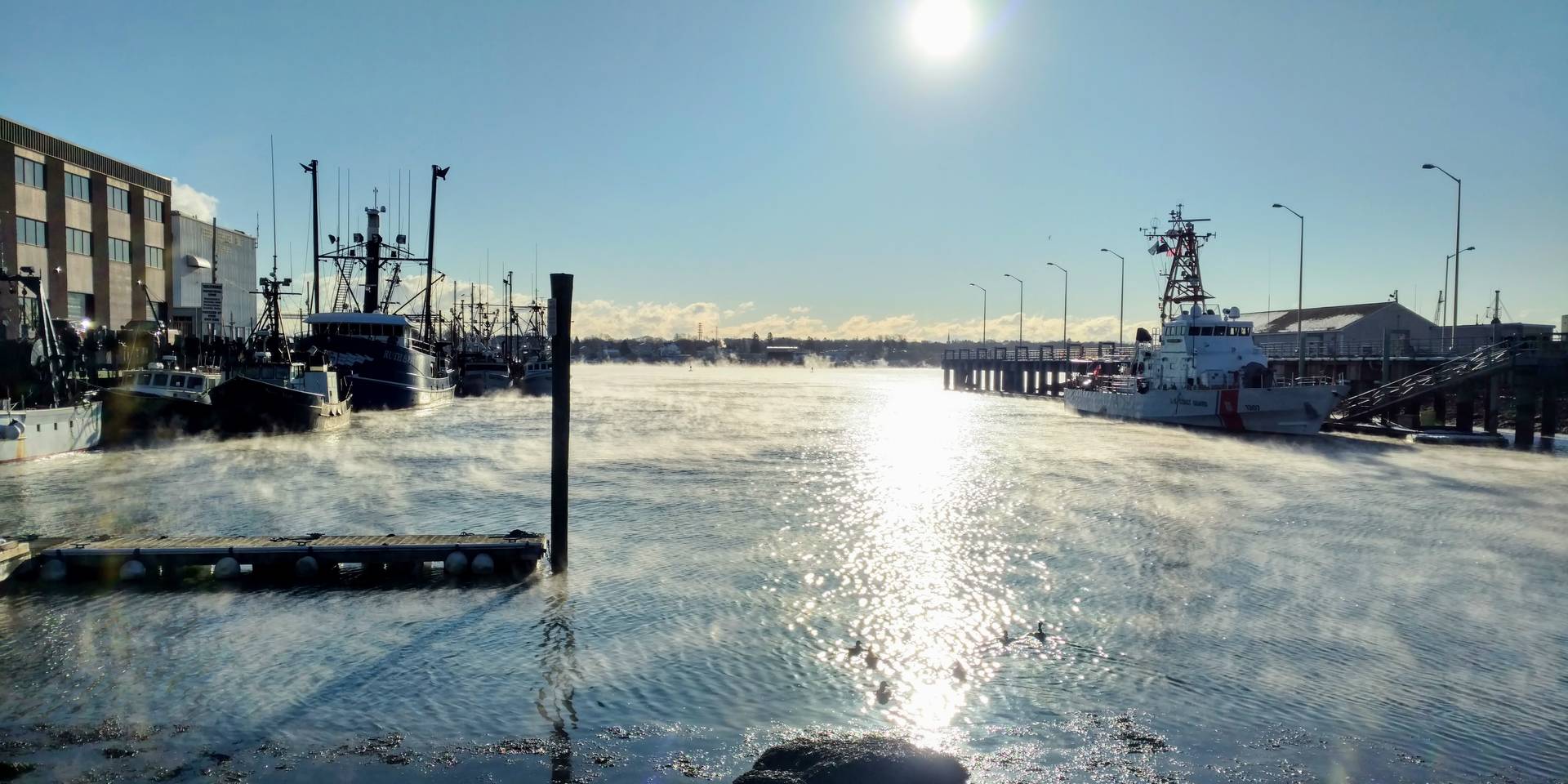 Early morning sun reflects on still water as fog lifts around boats at a working New England waterfront.